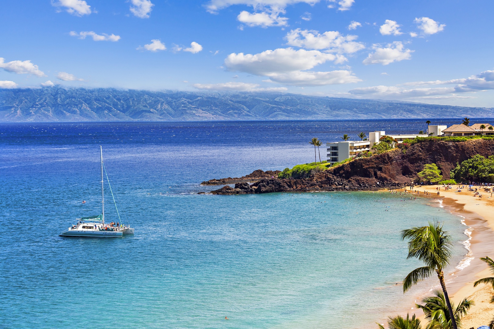 A stunning coastal setting where turquoise waters meet golden sand, with dramatic mountains rising beyond the deep blue ocean and a sailboat drifting peacefully offshore.