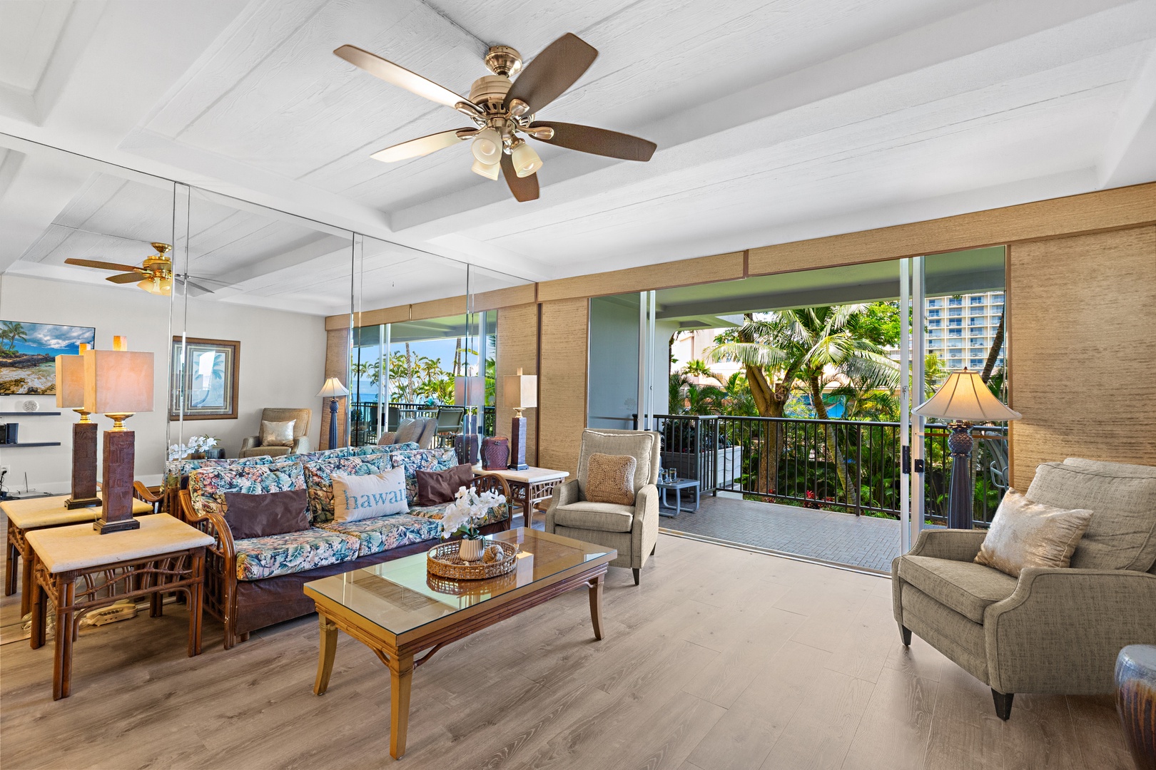 Relax in your bright tropical living room with ceiling fan, plush seating, and sliding doors opening to a balcony framed by lush greenery and ocean glimpses.
