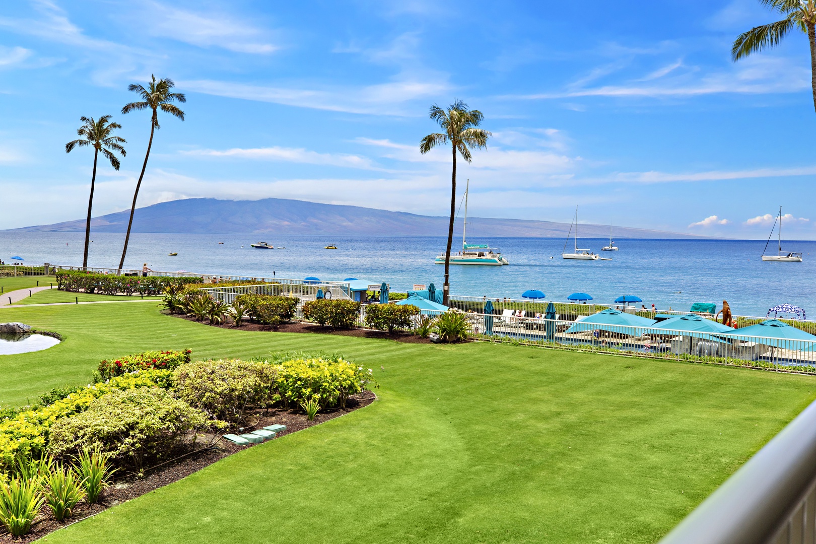 Stunning oceanfront property with manicured gardens, swaying palms, and pristine waters dotted with sailboats against a mountain backdrop.