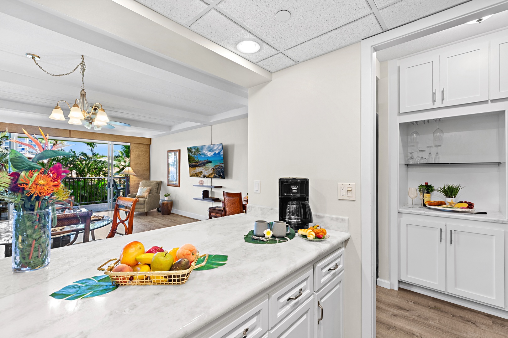 Step into your open kitchen featuring bright counters, fresh fruit accents, and a welcoming flow into your living space—where sunny balcony views and tropical touches await.