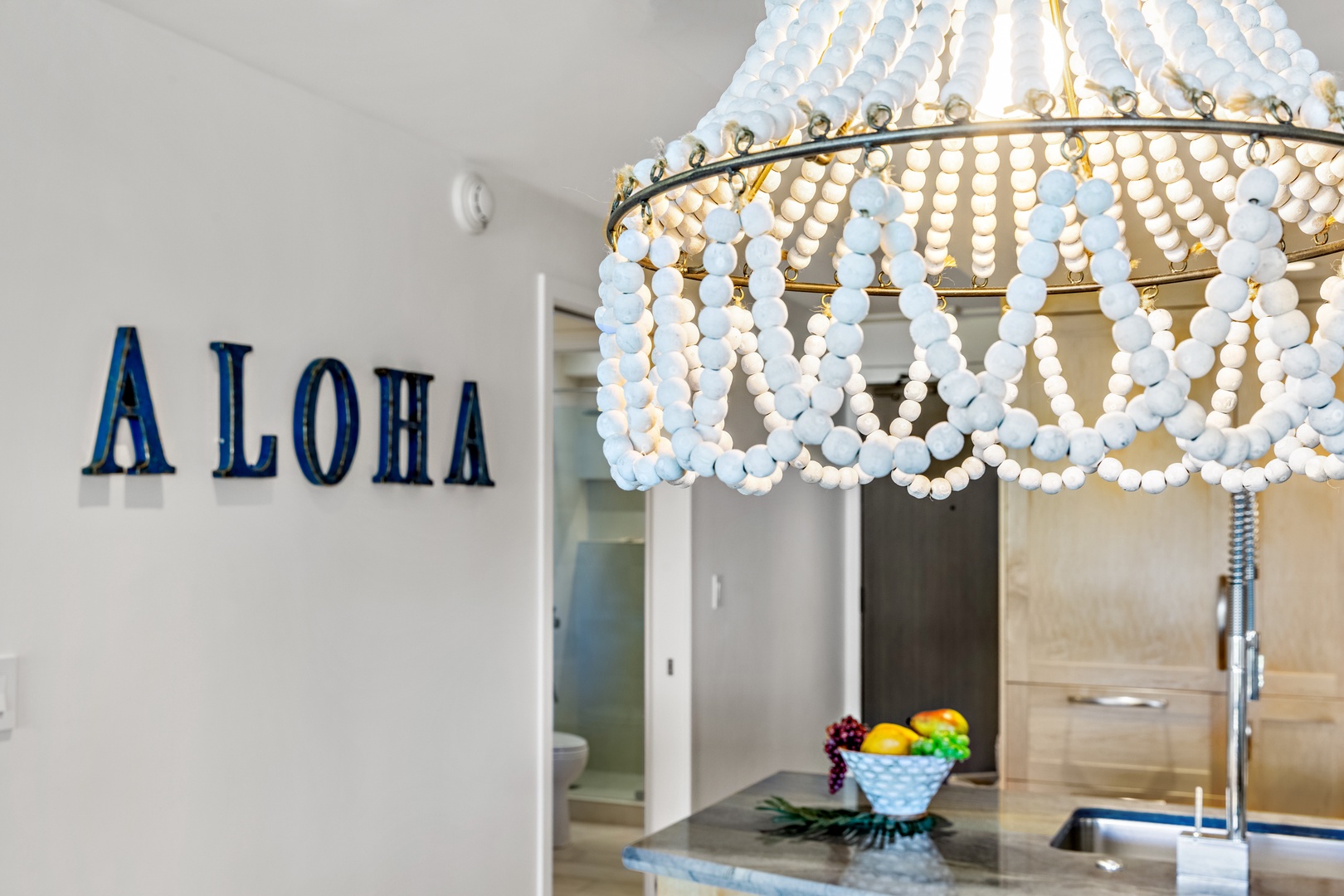 A welcoming Aloha greeting sets the tone as you step into the kitchen, lit by a stunning coastal-inspired beaded chandelier that casts a warm, inviting glow.
