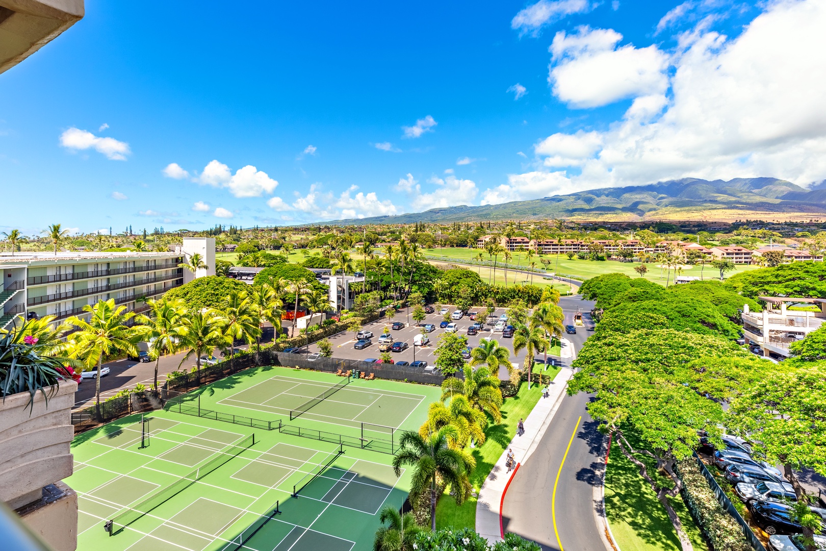 An expansive resort property featuring tennis courts, lush tropical landscaping, and striking mountain views, all set against a backdrop of blue Hawaiian skies.
