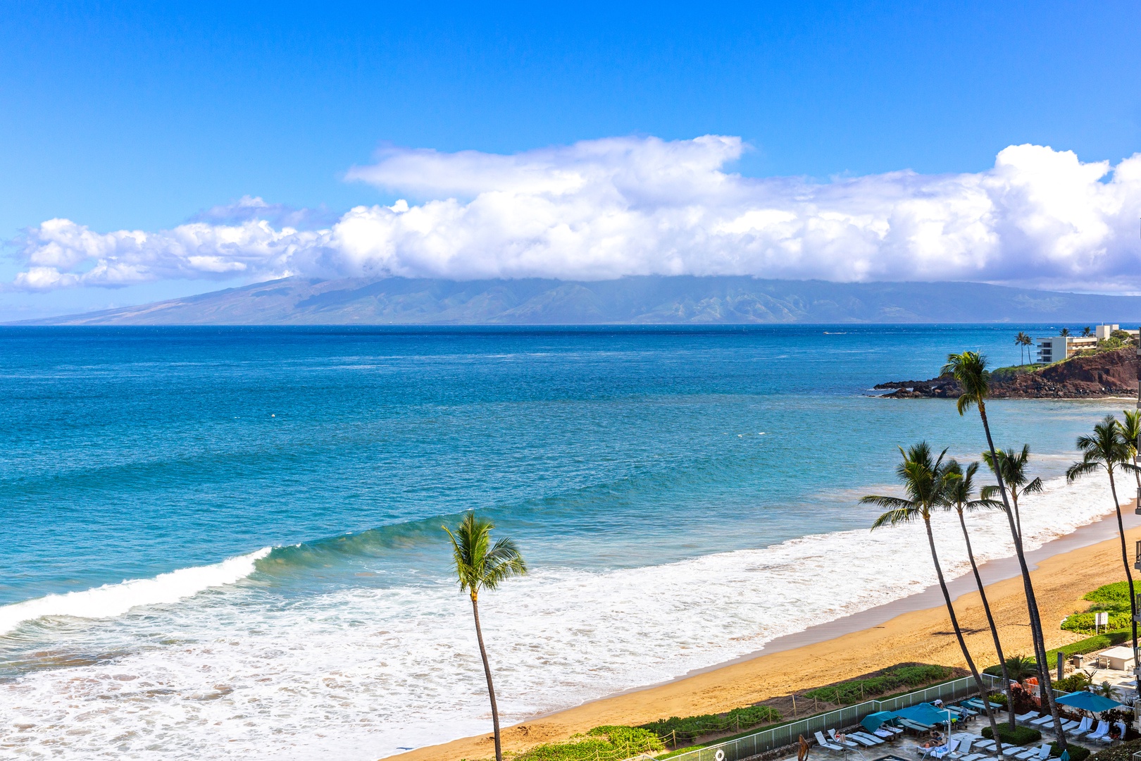 Stunning beachfront location with soft golden sands, swaying palms, and crystal-clear turquoise waters framed by distant mountain vistas and tropical skies.