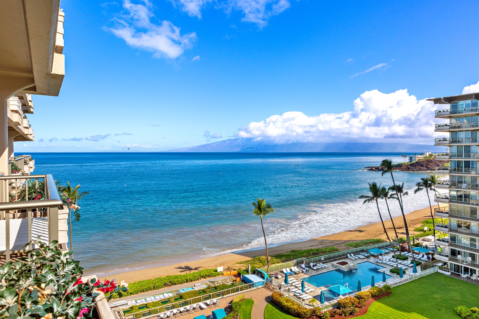 Oceanfront resort features a beachside pool and lush grounds with palms, overlooking turquoise waters and distant mountains under a bright Hawaiian sky.
