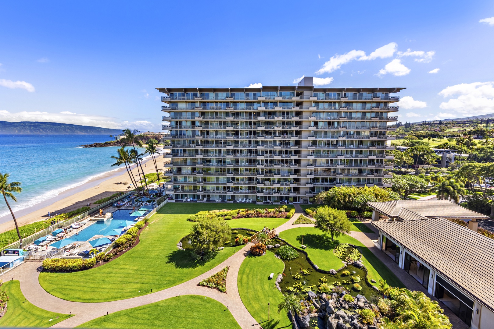 Manicured courtyard with tropical landscaping, winding pathways, and a resort-style pool framed by oceanfront buildings.