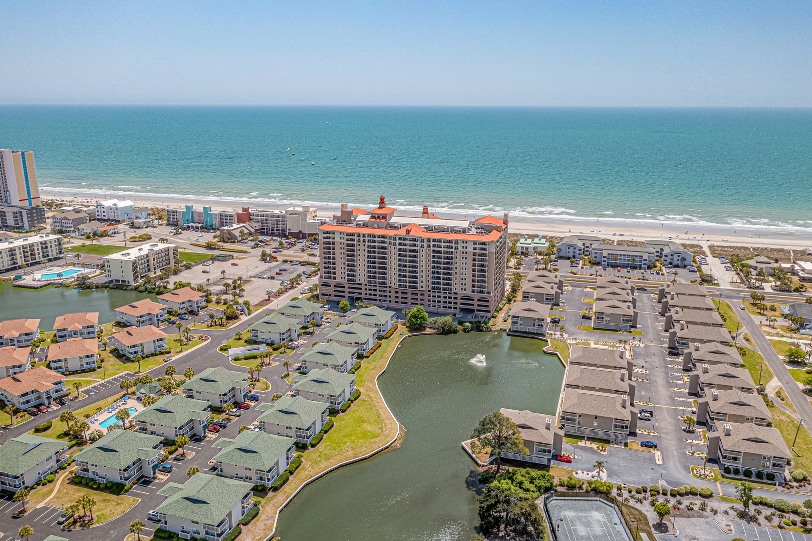 Aerial view of a beachfront resort community with a central pond, featuring colorful buildings and direct access to the sandy shore and sparkling ocean.