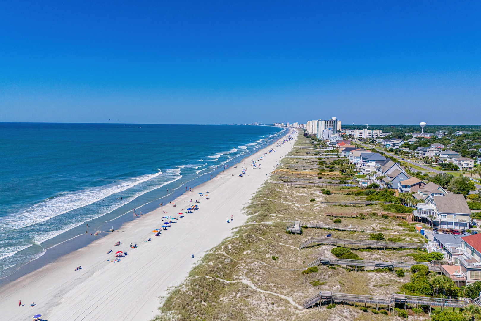 An aerial view showcasing the pristine coastal area with expansive sandy shores, rolling waves, and residential buildings nestled along the shoreline under a clear blue sky.