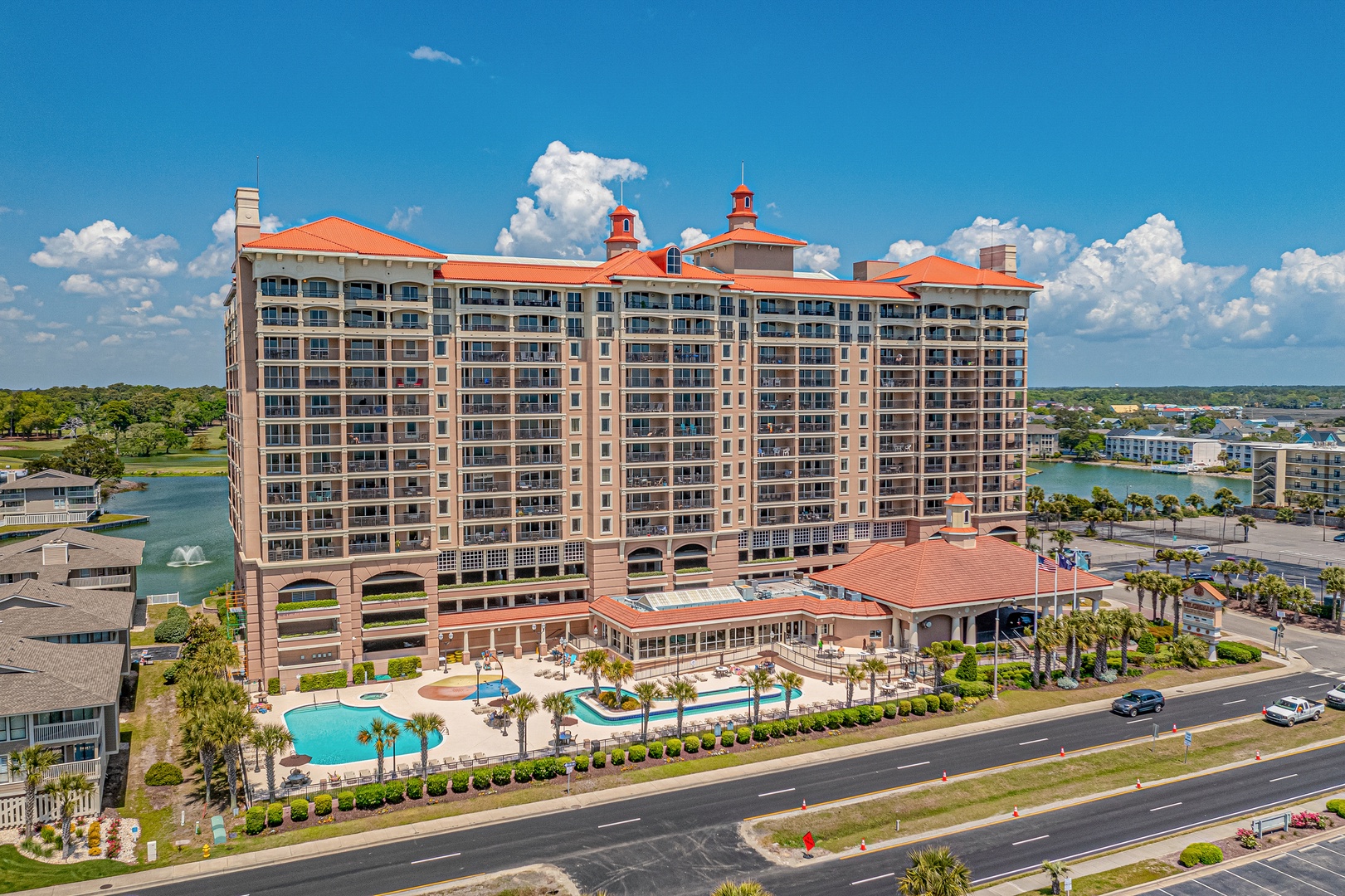A grand waterfront resort featuring Mediterranean architecture with terracotta roofs, multiple balconies, and a sparkling pool surrounded by palm trees under sunny skies.