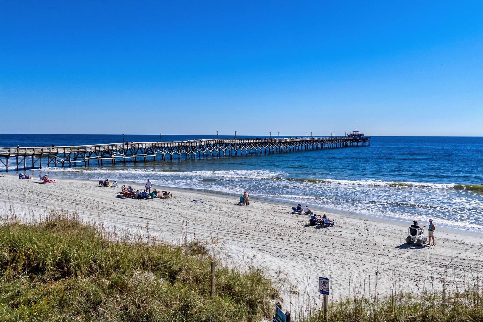 A pristine sandy beach stretches along the Atlantic coast with a historic wooden pier extending into the sparkling blue waters—a landmark feature of the surrounding area.