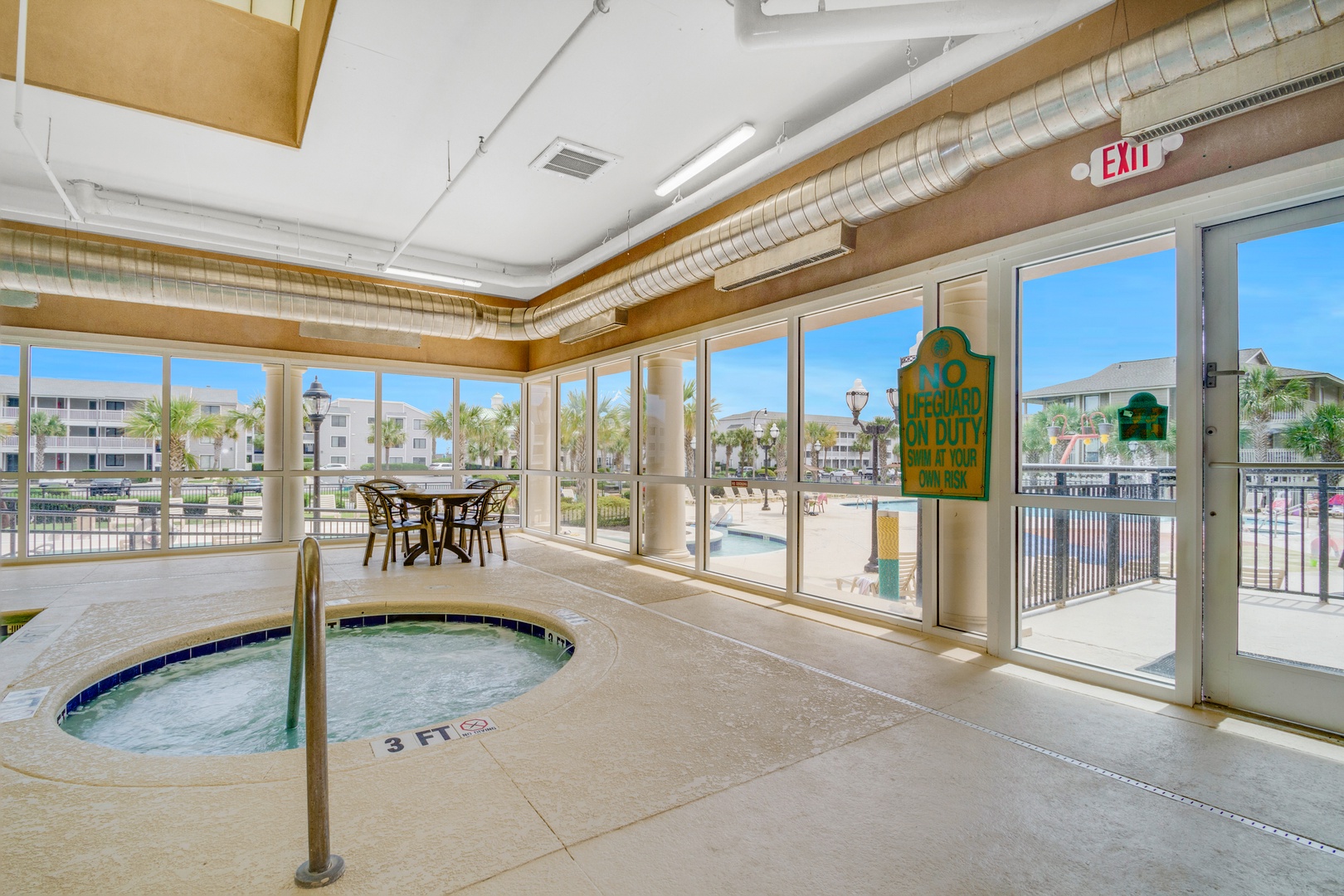 Indoor pool area with hot tub and tropical views through floor-to-ceiling windows. The seating area leads to an outdoor deck with palm trees and sunshine.
