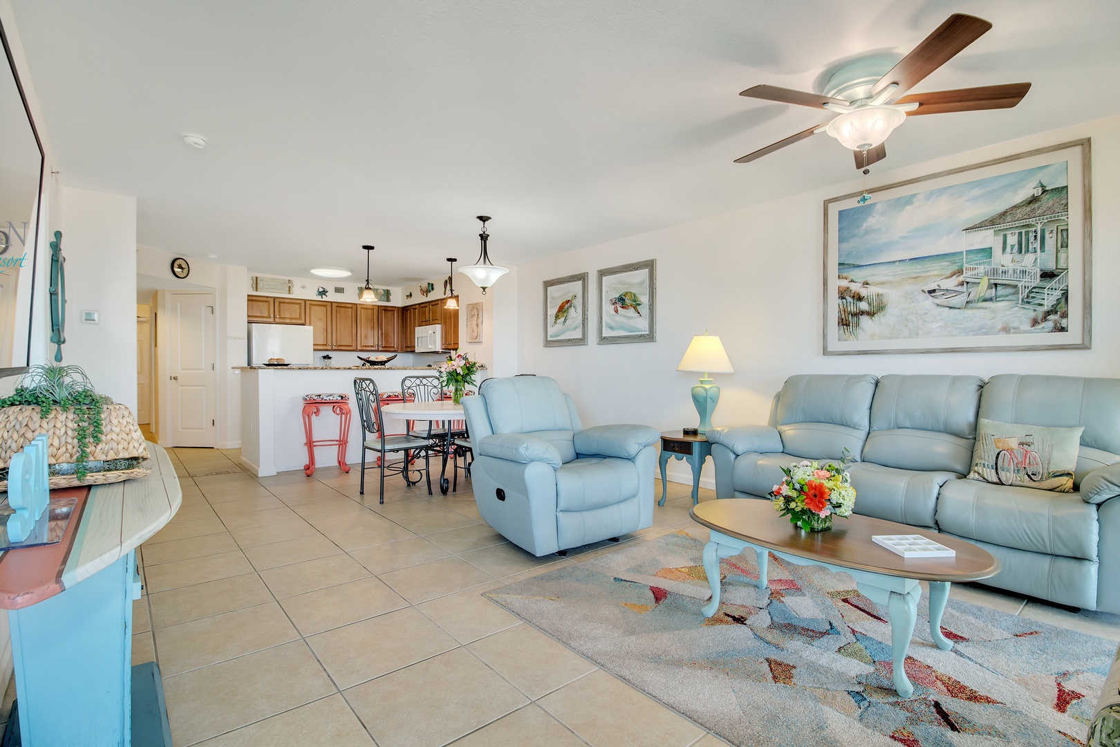 Relax in your spacious coastal living room with soft blue furnishings, ceiling fan, and open kitchen—perfect for unwinding after beach days.