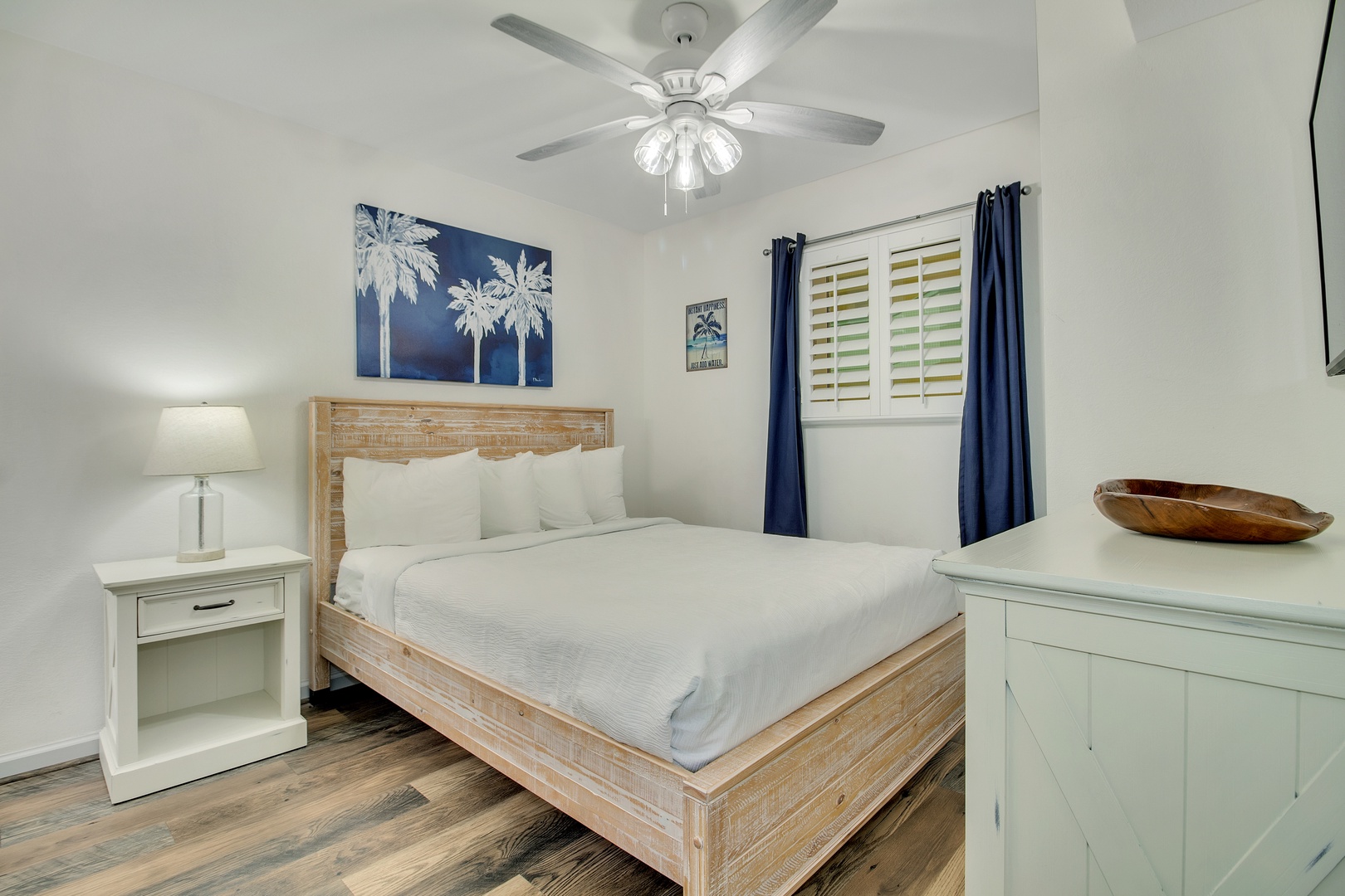Unwind in your serene bedroom featuring crisp white linens, coastal palm artwork, and gentle ceiling fan breezes for perfect comfort.