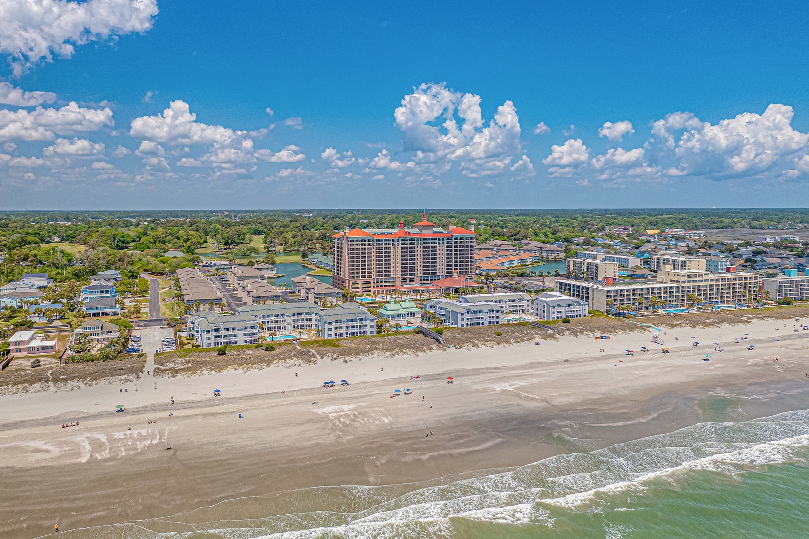 Expansive beachfront community with coastal residences and resort buildings stretching along a pristine sandy shore, surrounded by lush greenery and open skies.