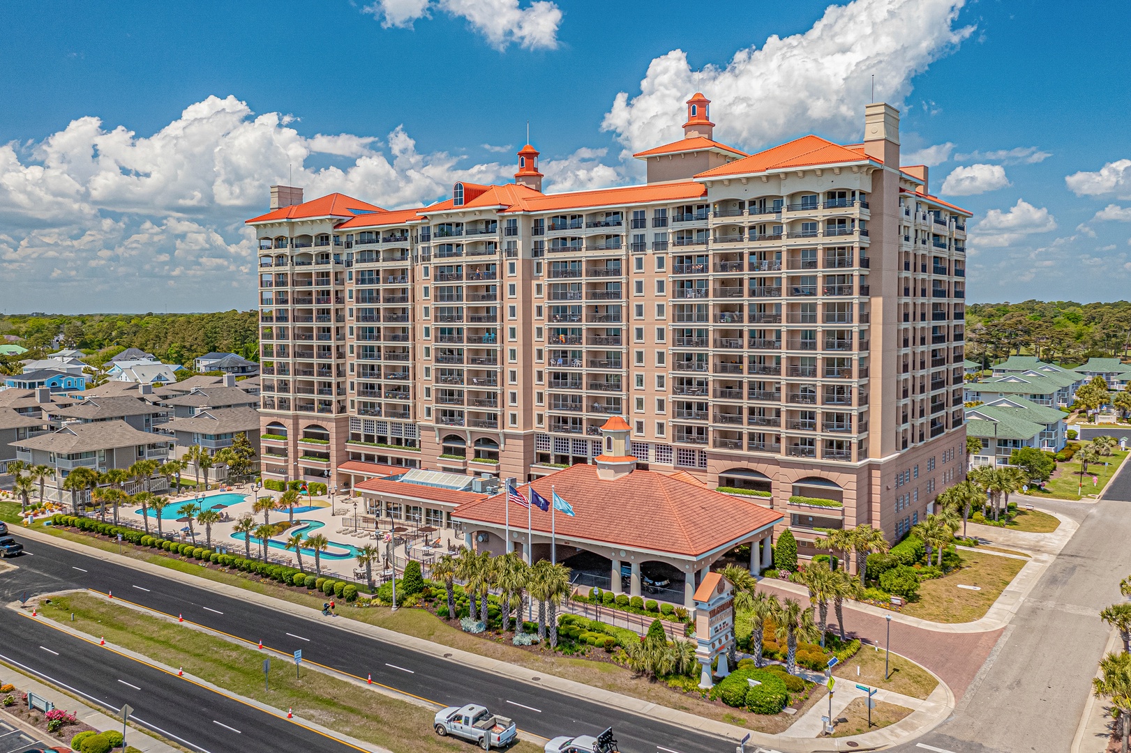 Elegant Mediterranean-style resort building with distinctive terracotta roofing, pool facilities, and palm-lined entrance in a coastal setting surrounded by vacation homes and green spaces.