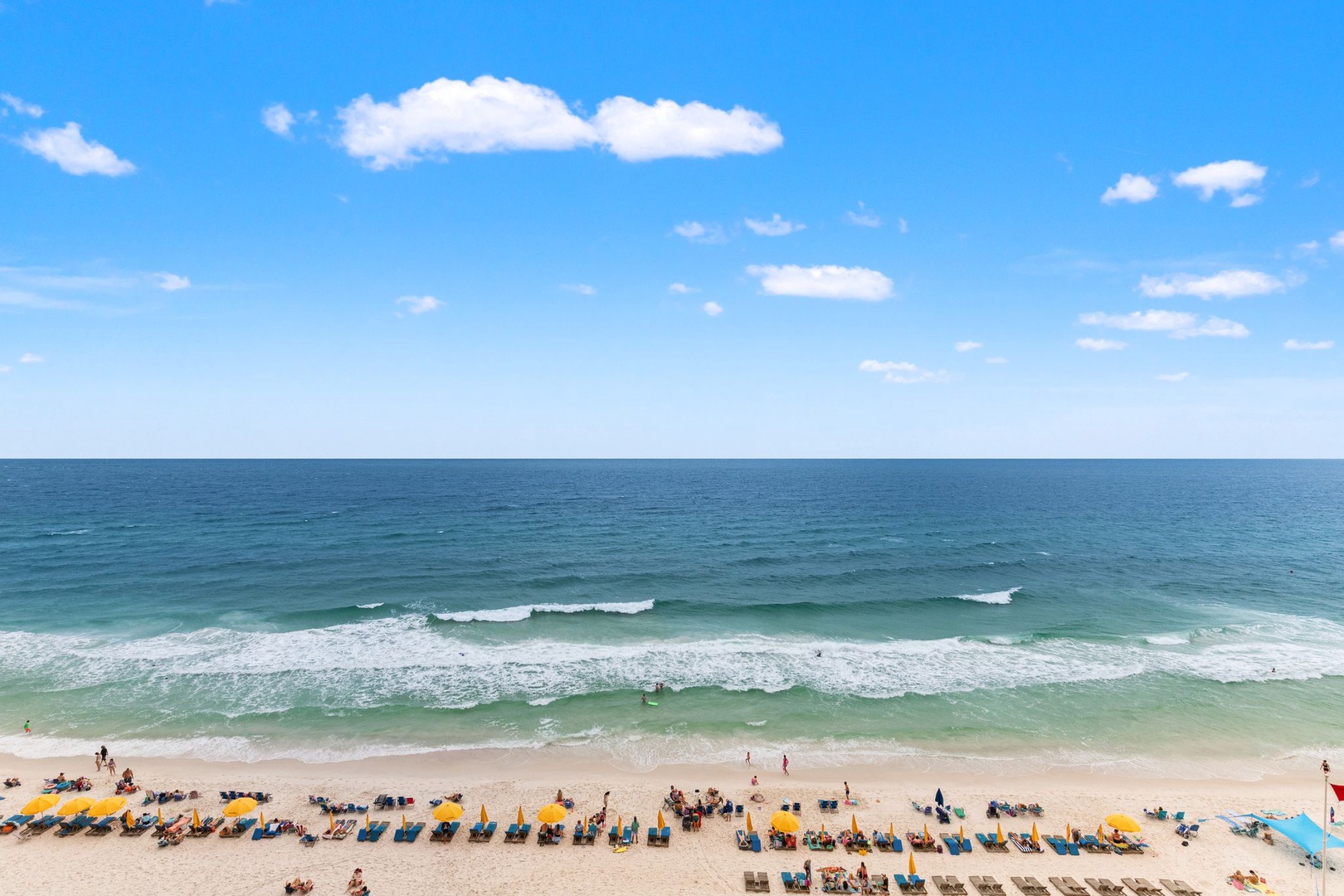 Aerial view of a pristine beach with clear turquoise waters and rows of sunbeds lining the golden sand under a bright blue sky.