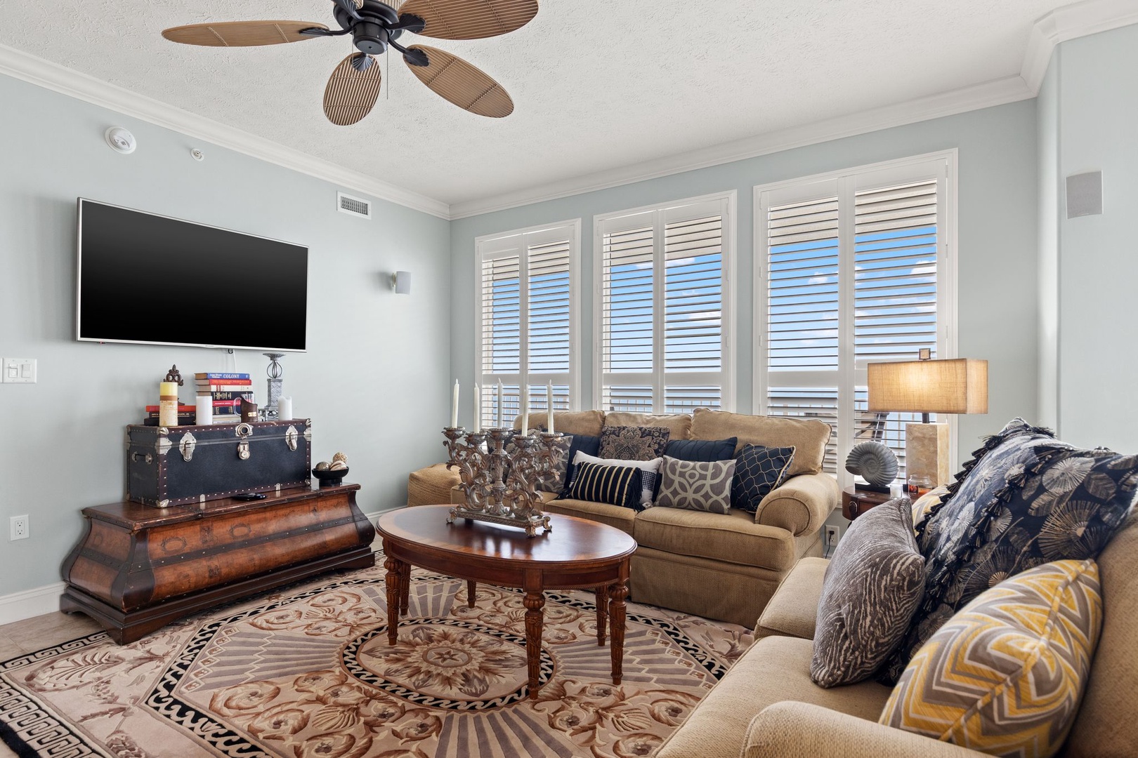 Your comfortable living room features plush seating, a wall-mounted TV, and coastal-inspired décor. Elegant shutters frame bright windows, while a ceiling fan keeps the space refreshingly cool.