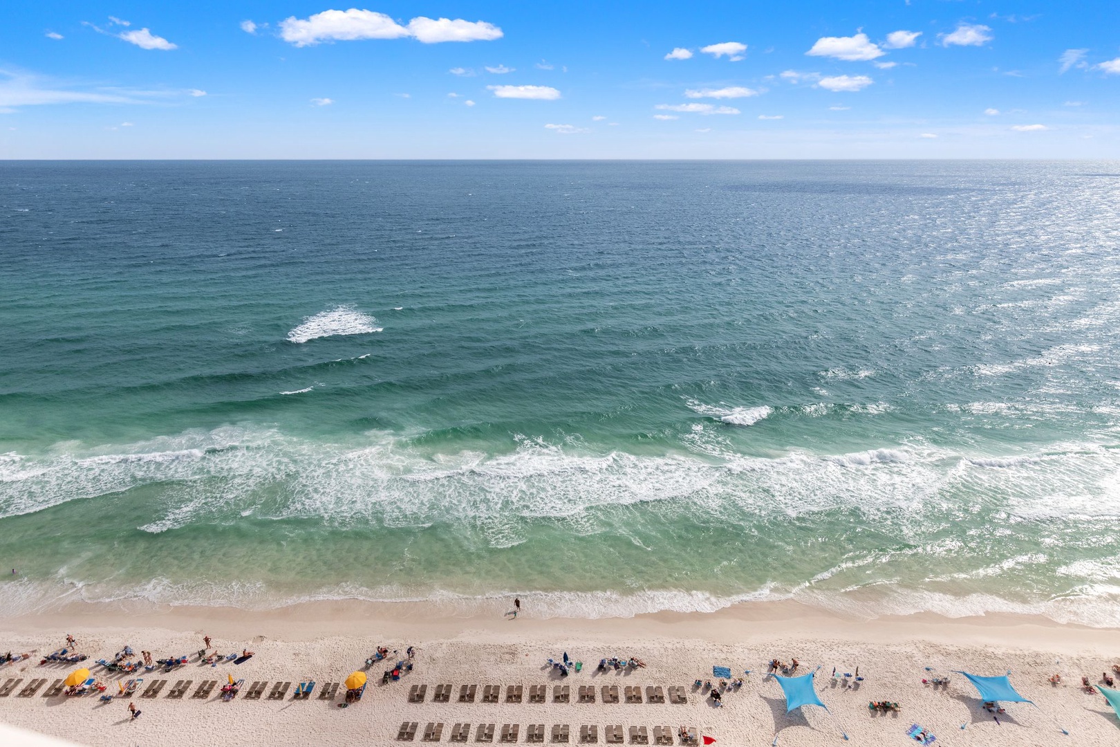 Aerial perspective showcasing the property's beachfront location with pristine emerald waters, white sand beach, and organized sun loungers. The view captures the natural beauty of the Gulf coast setting and surrounding beach amenities.