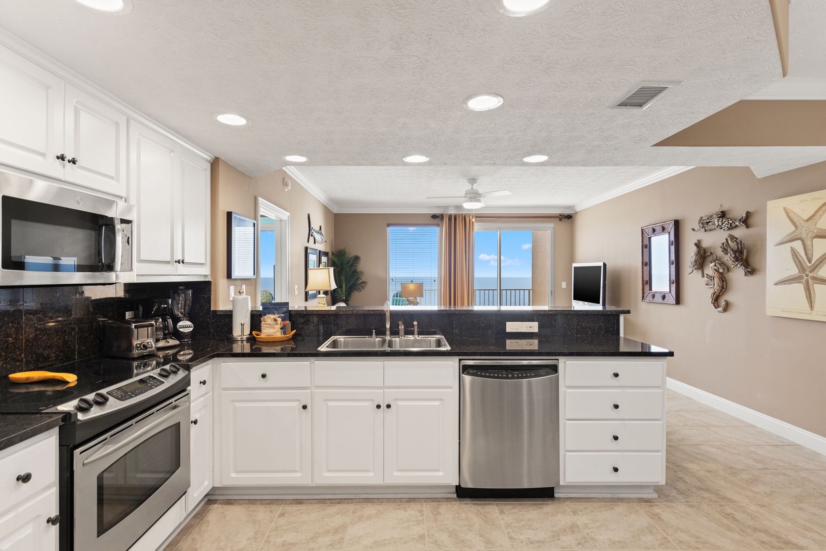 Your spacious kitchen features modern stainless appliances, elegant dark granite counters, and bright white cabinetry—all set against coastal-inspired décor and stunning sea views through the windows.