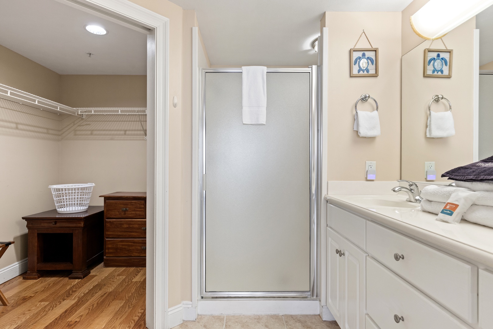 Your private bathroom offers fresh towels, a glass-enclosed shower, and a spacious vanity with coastal turtle artwork—plus a walk-in closet with ample storage for all your belongings.