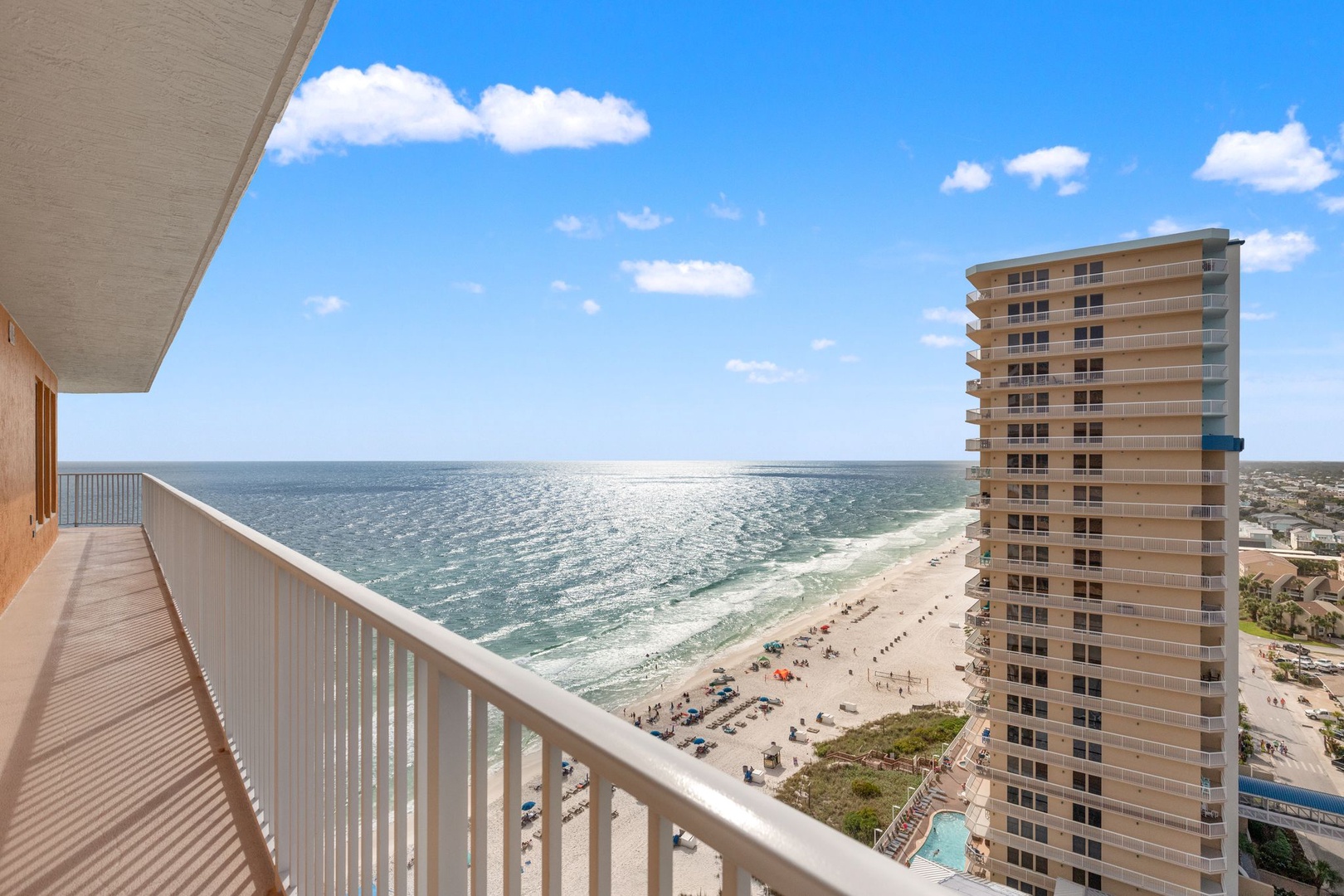 A spacious beachfront balcony with expansive ocean views, white sand beaches, and nearby high-rise buildings in a sunny coastal setting.