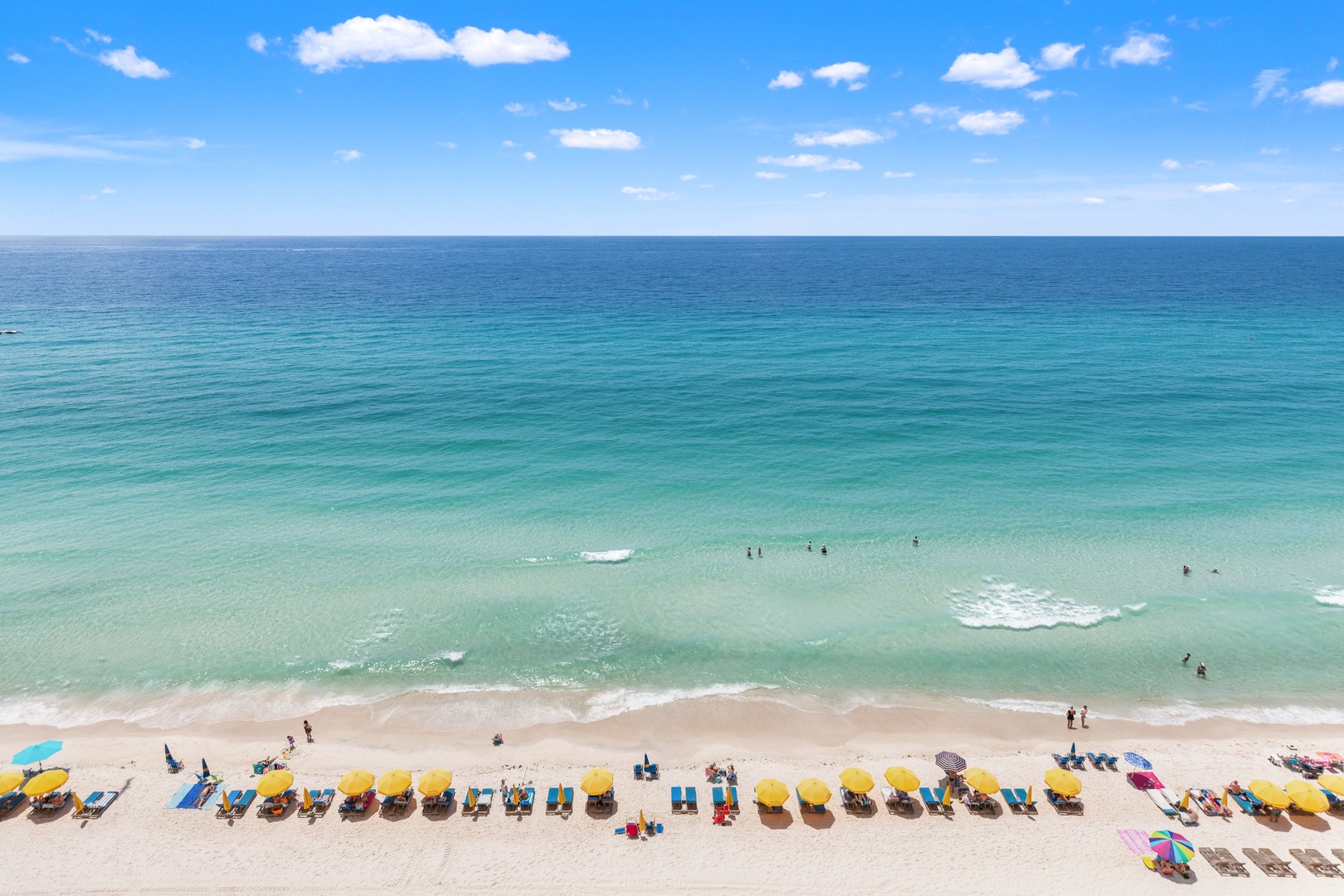 Stunning beachfront location with pristine turquoise waters and soft white sand, complemented by rows of colorful umbrellas and lounge chairs along the shore.