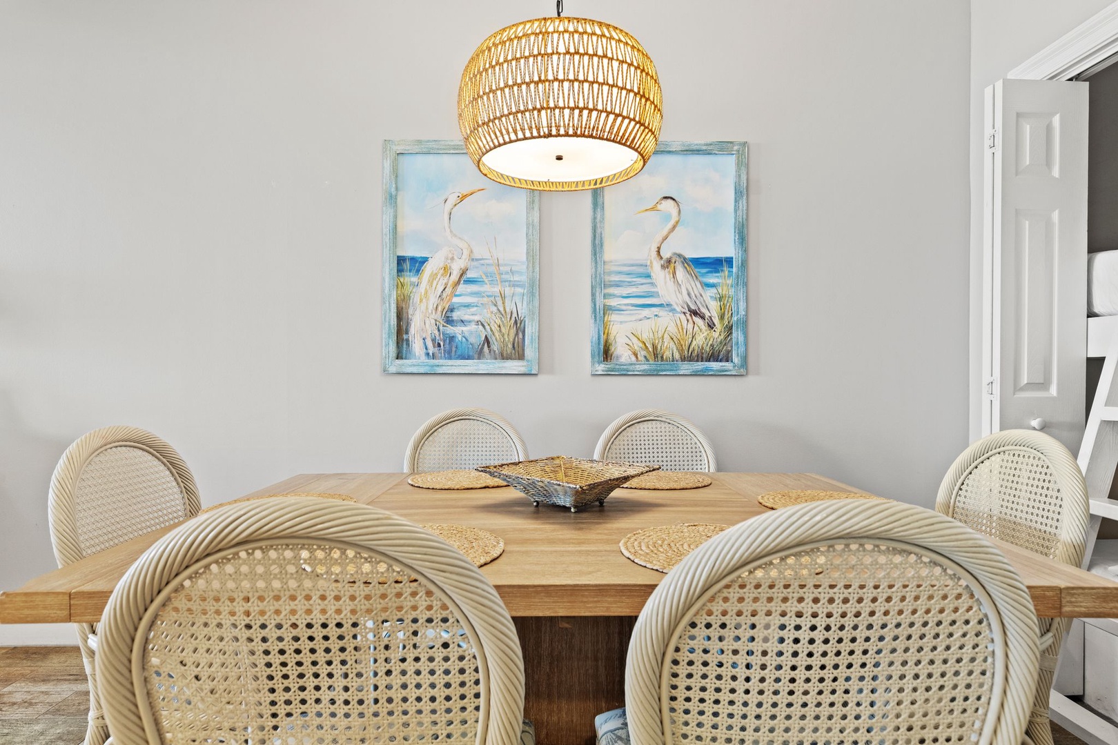 A light-filled dining space with coastal artwork, a woven pendant, and a natural wood table ready for shared meals and conversation.