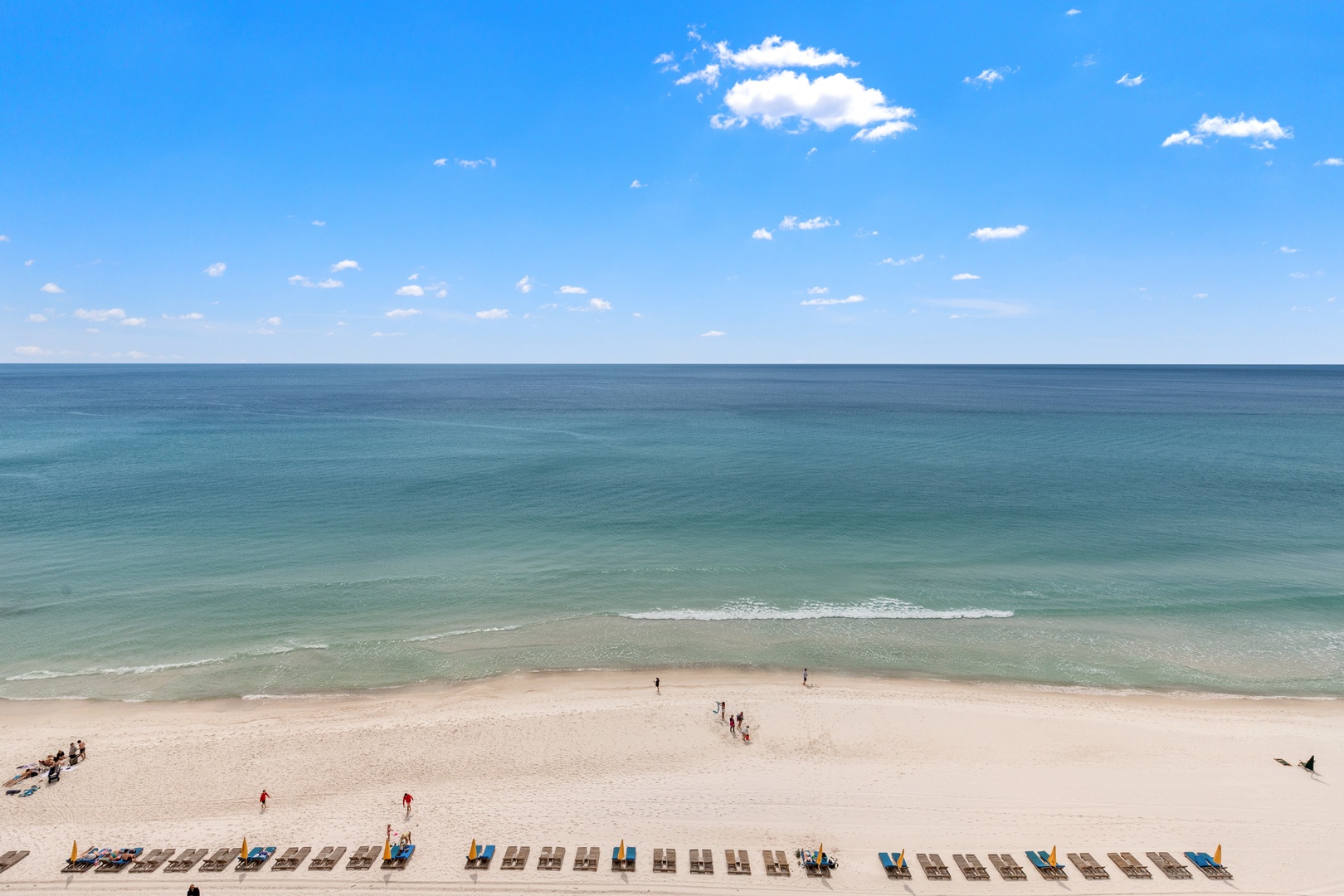 Pristine white sand beach stretches along turquoise waters under endless blue skies with scattered beachgoers enjoying the coastline.