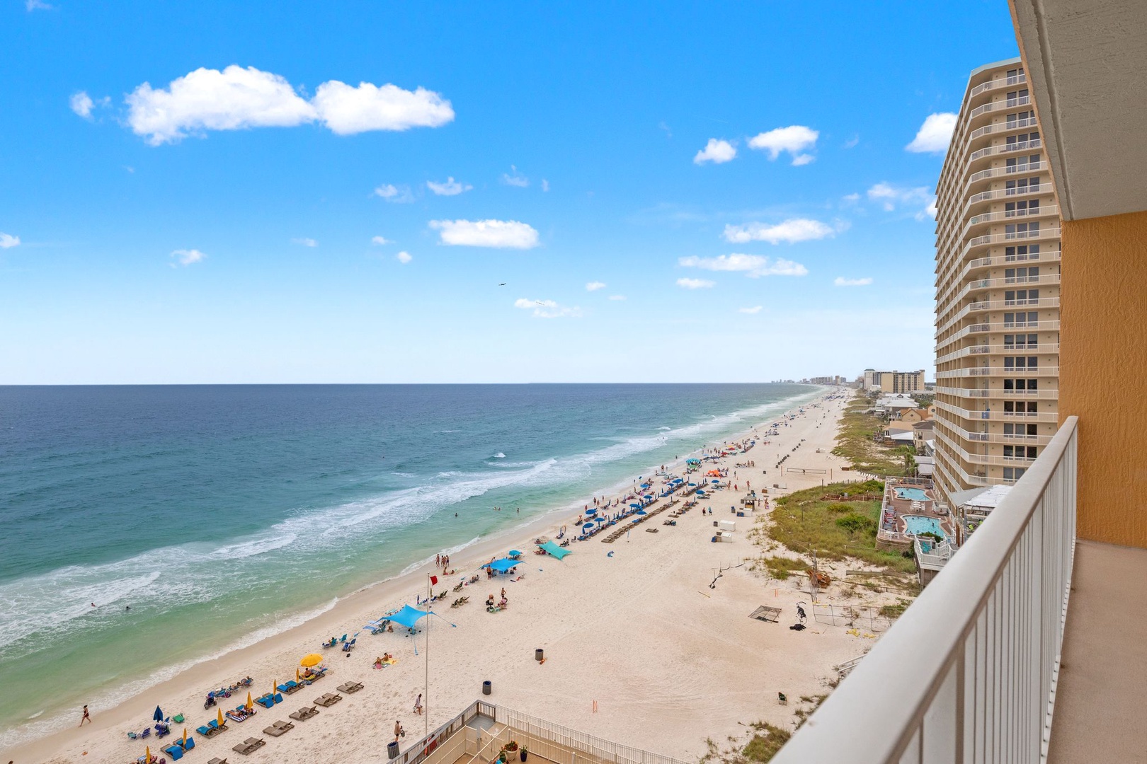 This view captures the property's beachfront location along a pristine stretch of white sand, with neighboring buildings and the Gulf Coast shoreline extending into the distance under clear blue skies.