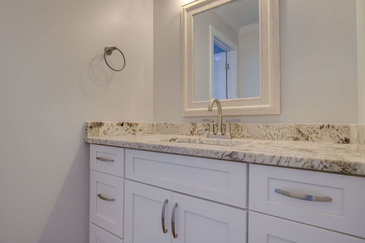 A bathroom with a marble countertop, a sink with a faucet, and a mirror with a decorative frame. There are white cabinets with silver handles.