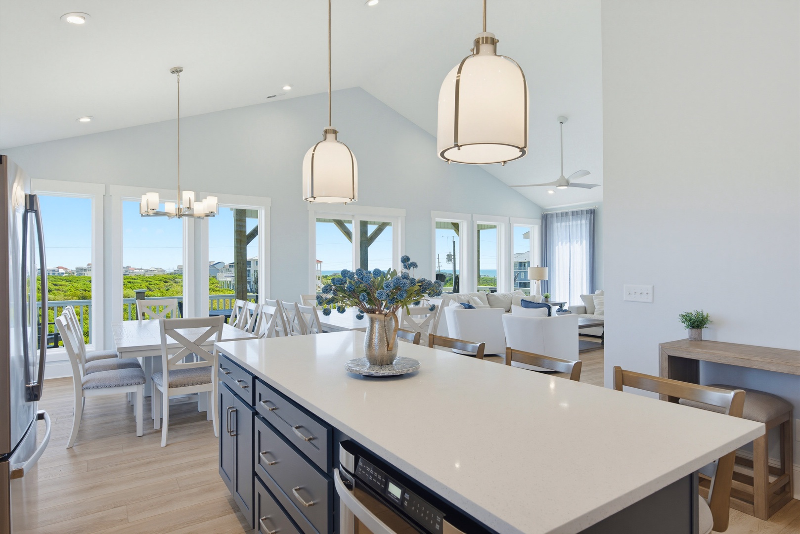 Bright coastal great room with vaulted ceilings, elegant pendant lights, and a fully equipped kitchen island. Expansive windows frame seamless dining and living spaces flooded with natural light.