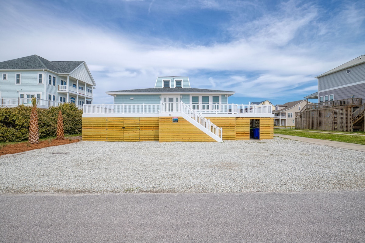 A beachfront property with a wooden deck, a blue house, and a gravel driveway.