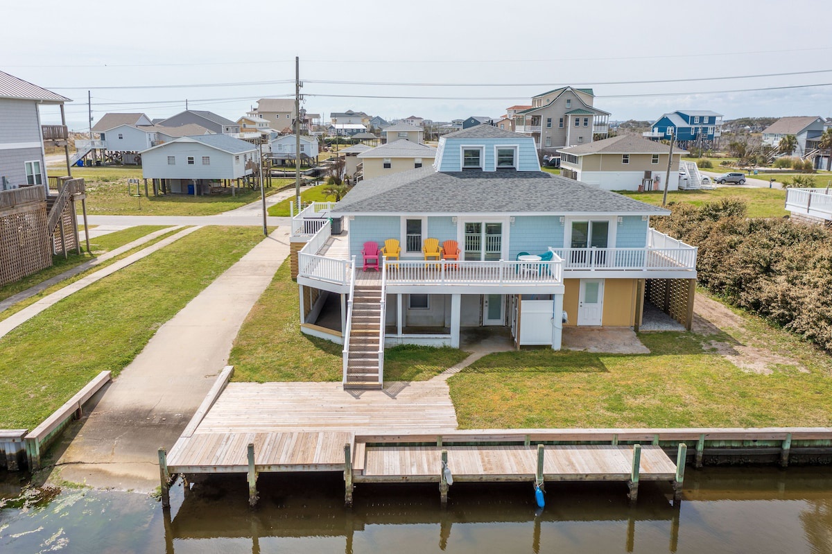 A two-story house with a dock extending into a body of water, surrounded by a grassy area and other houses.