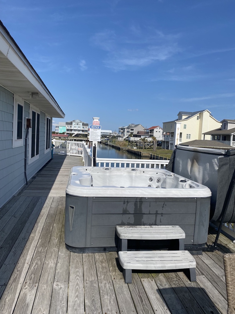 A wooden deck with a hot tub and a bench, overlooking a water body and residential buildings.