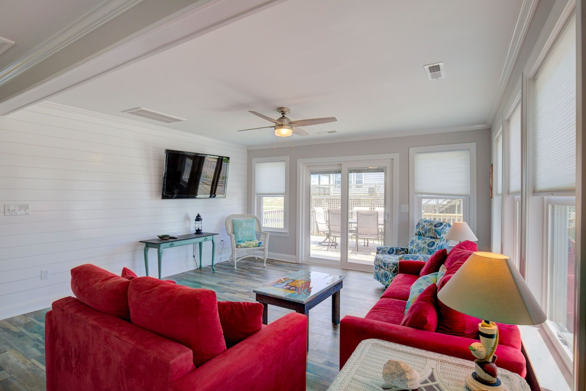 A spacious living room with a red sofa, a coffee table, and a ceiling fan. There is a TV mounted on the wall and a window with blinds.