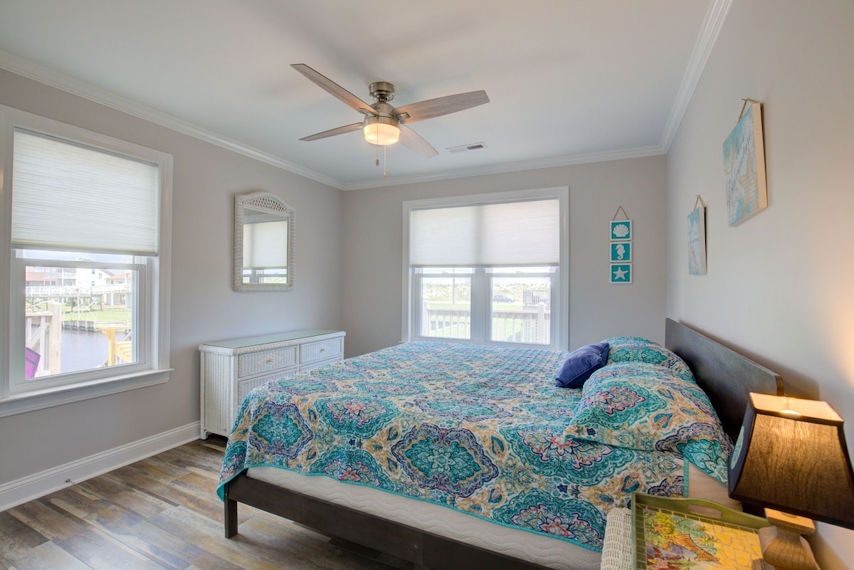 A bedroom with a large bed, a ceiling fan, a window with blinds, a radiator, and a wall-mounted shelf.