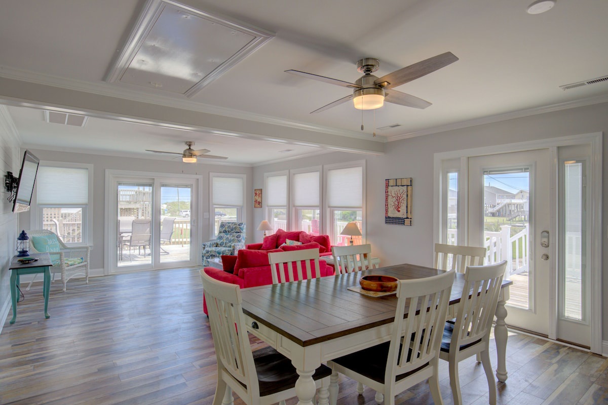 A spacious dining area with a wooden table and white chairs, a bright ceiling fan, and large windows offering a view of the outdoors.