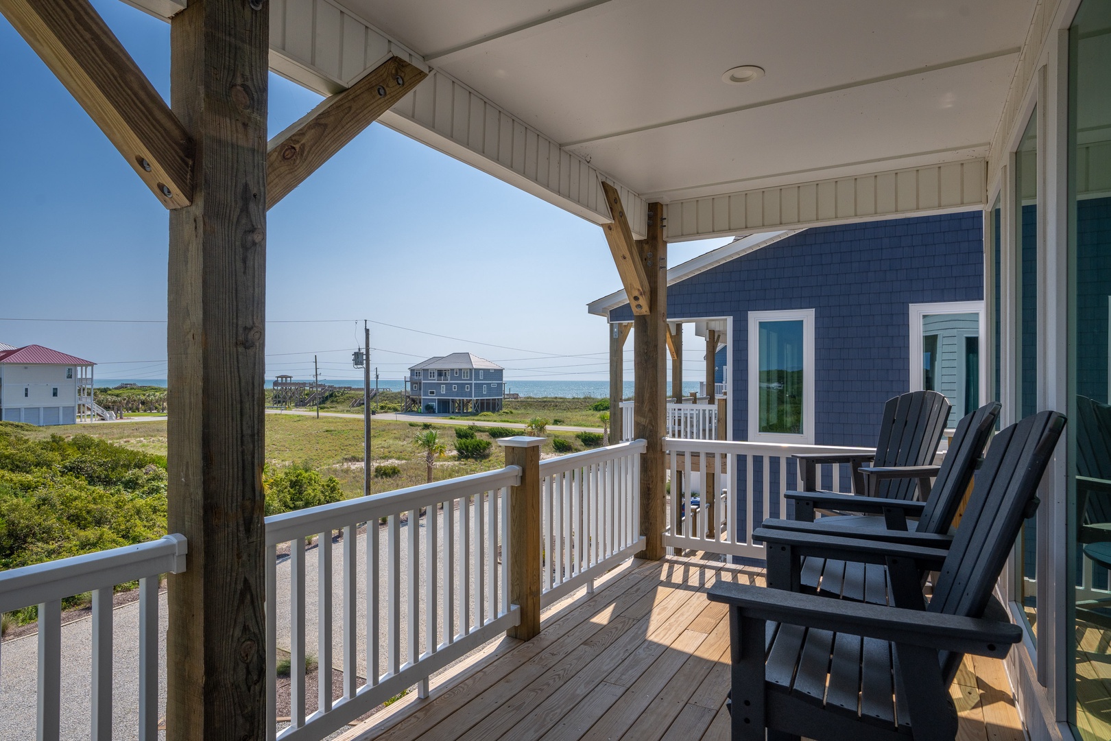 A covered porch with Adirondack chairs frames coastal views across dunes and neighboring beach houses. Settle in with your morning coffee and soak in the ocean breeze.