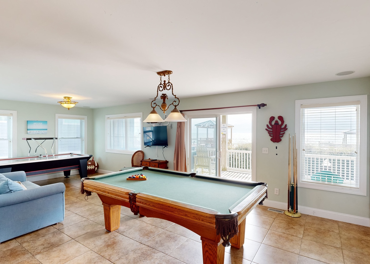 Your game room invites fun with a classic pool table, air hockey, and coastal charm. Large windows and patio doors let in natural light while framing the outdoor deck beyond.