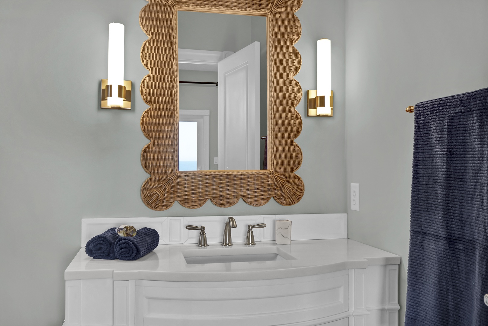 Step into a coastal-inspired bathroom where a stunning woven mirror and soft gray walls create your serene retreat. Luxurious towels await, perfectly coordinated to make you feel pampered and relaxed.