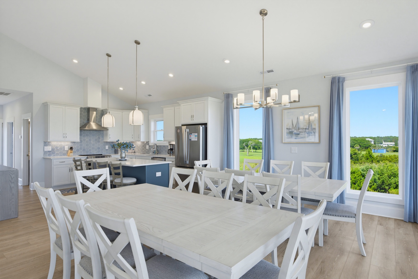 Bright, open-concept living space with expansive dining table and floor-to-ceiling windows overlooking lush greenery. Modern kitchen perfect for cooking and gathering together.