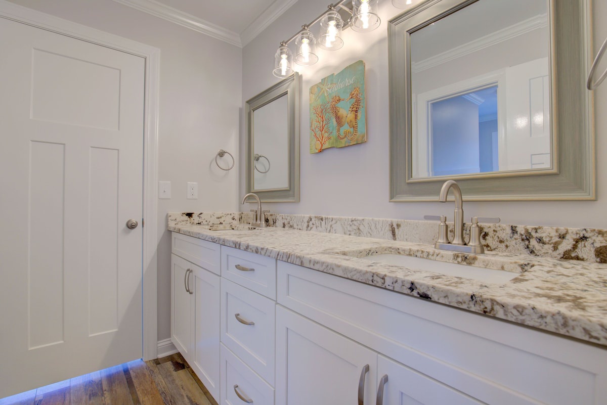 A bathroom with white cabinetry, a marble countertop, and a large mirror with lights above.