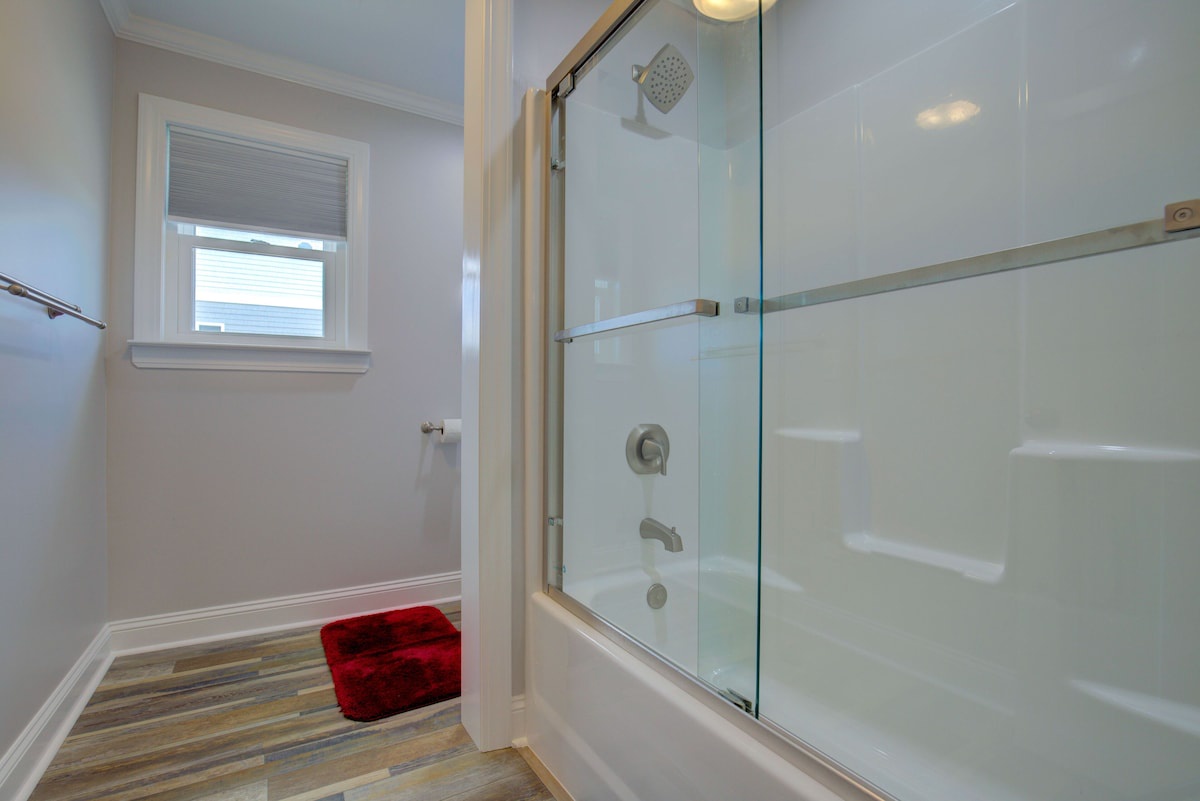 A modern bathroom with a glass shower enclosure, a wooden floor, and a red bath mat.