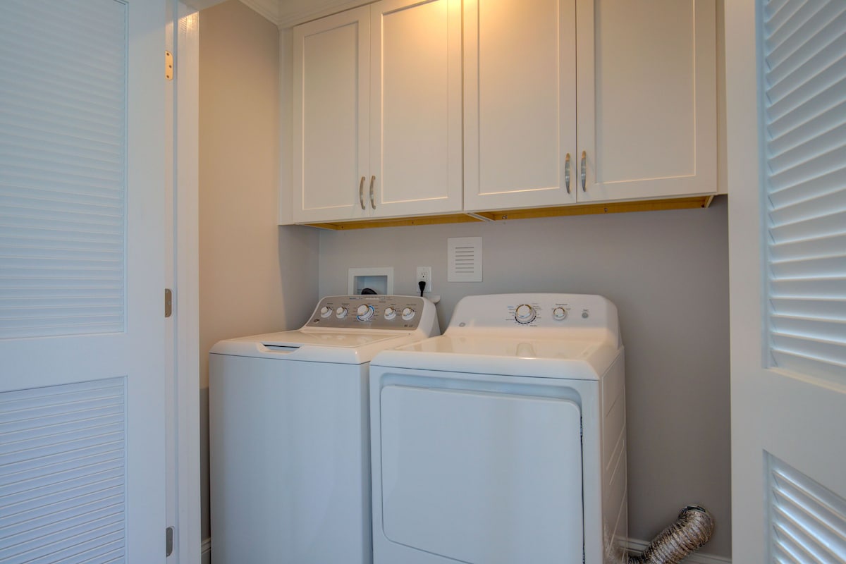 A laundry room with two white front-loading washers and dryers, white cabinets above, and a vent hose on the right.