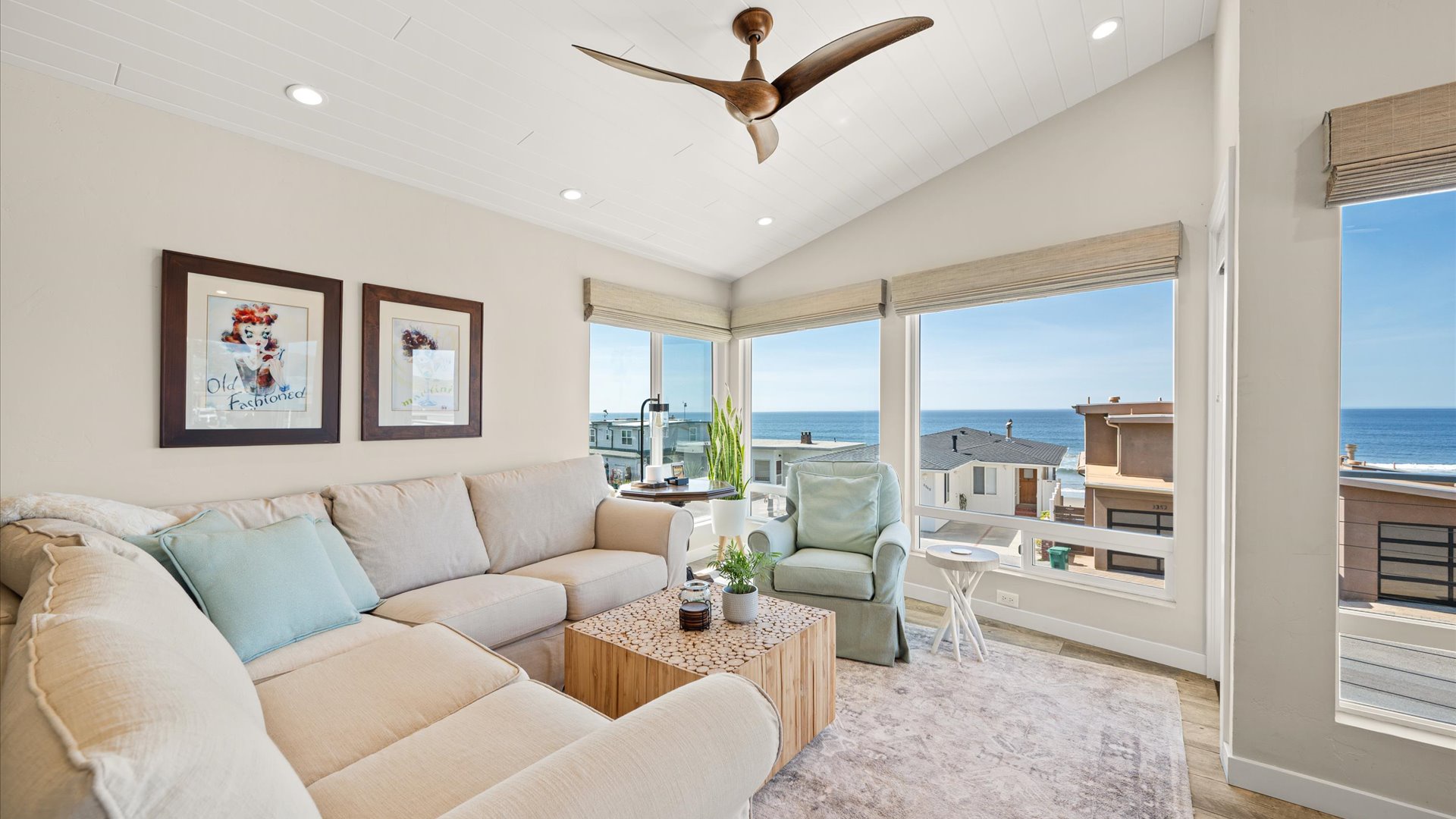 The bright living room features ocean views through wall-to-wall windows, comfy seating, and a gentle ceiling fan for perfect coastal relaxation.