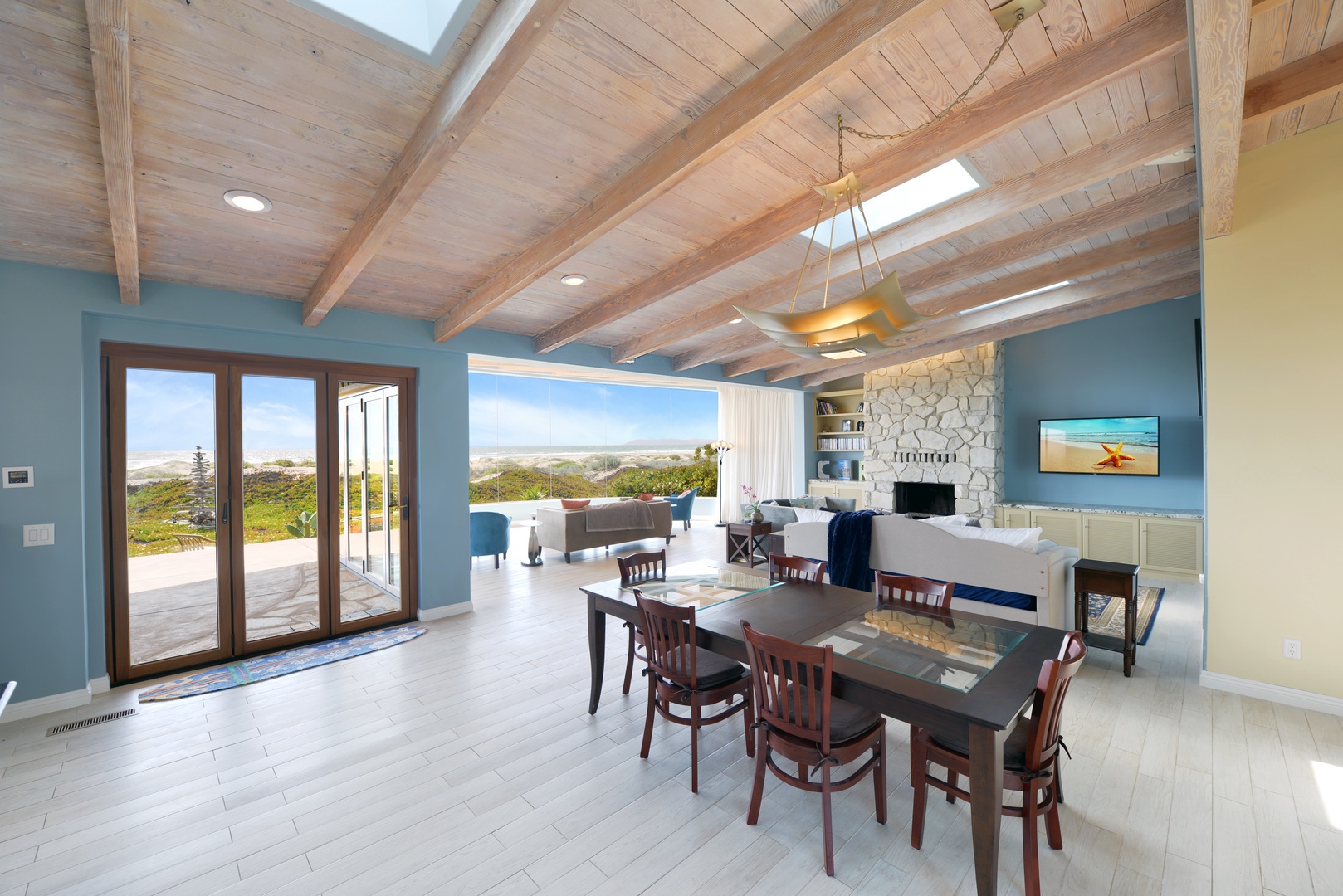 Open-concept retreat with rustic wood beams, skylights, and glass doors framing sweeping coastal views. Gather at the dining table or unwind by the stone fireplace.