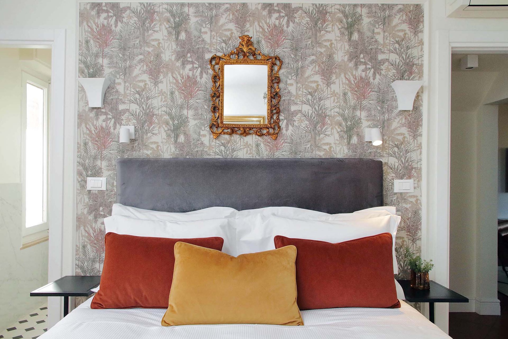 Sink into your plush bed adorned with vibrant pillows, where elegant botanical wallpaper and ornate touches create a perfect sanctuary for rest.