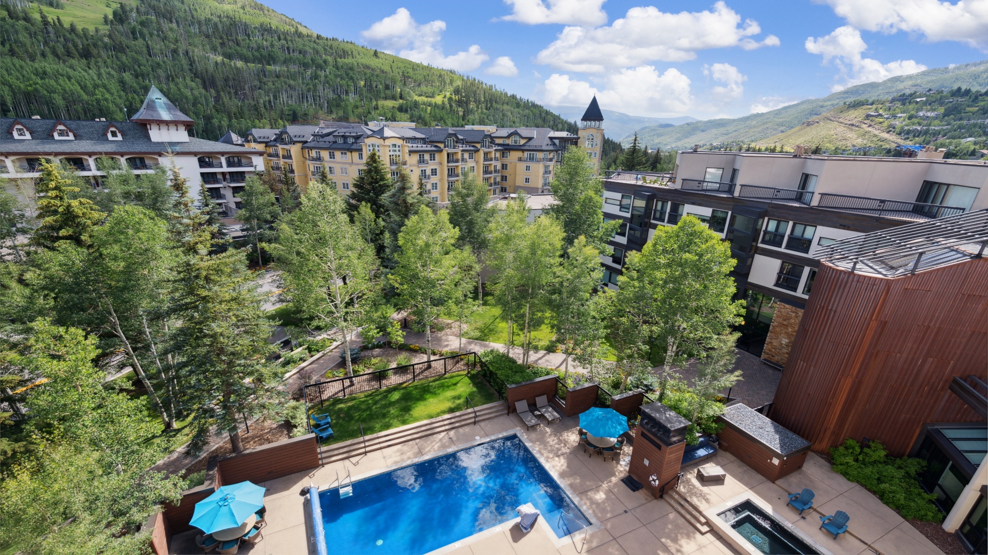 A mountain resort nestled in a lush valley, featuring a central pool area surrounded by tree-lined grounds and multi-level lodges with scenic alpine slopes in the distance.