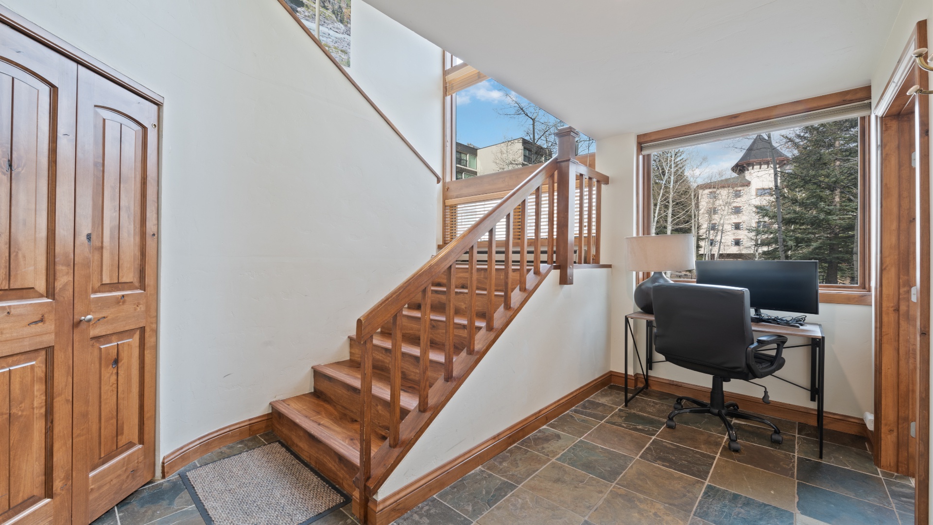 Step into your bright mountain retreat where a warm wooden staircase leads to upper floors, and a handy workspace awaits by windows framing snowy peaks and evergreens.