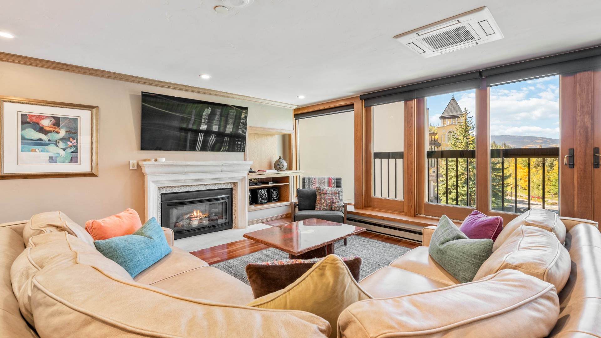 Curl up on plush sofas by the fireplace and enjoy mountain views through large windows—your cozy retreat awaits with vibrant pillows and a peaceful ambiance.