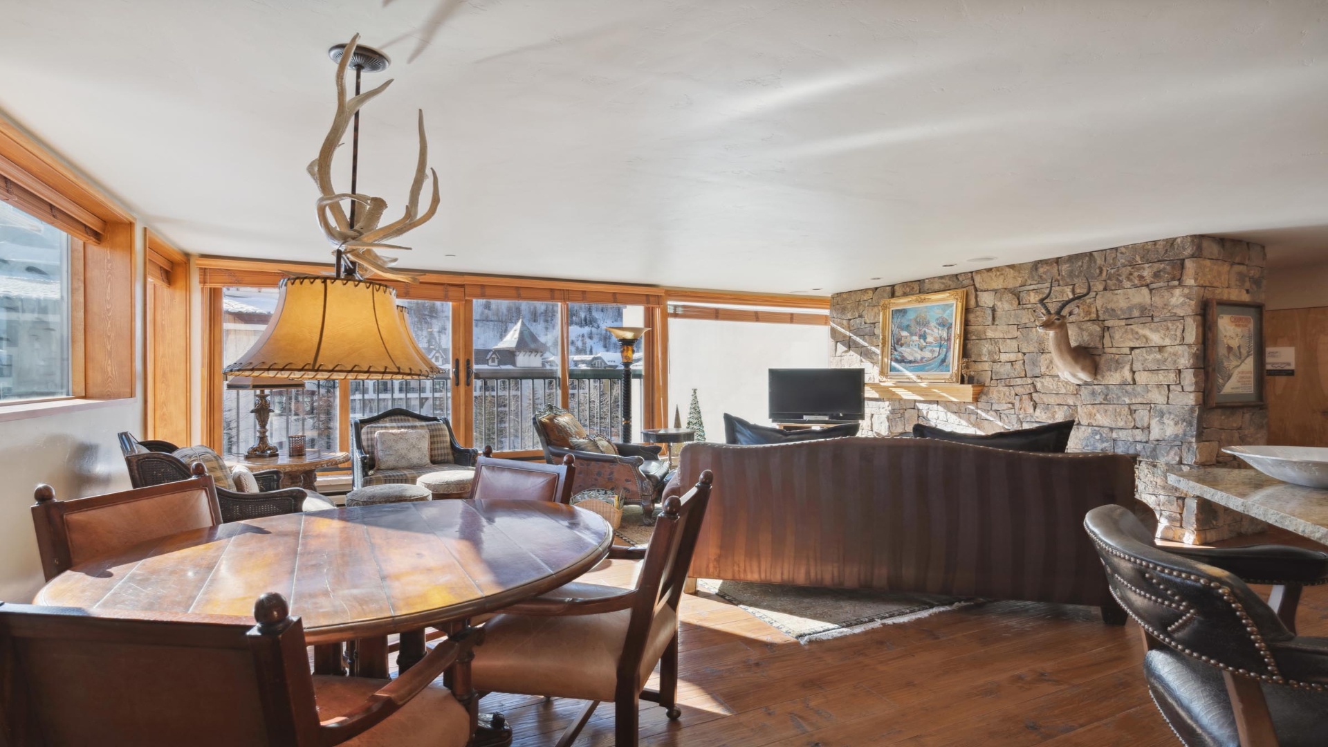 Relax in a spacious, sunlit living area featuring rustic elegance with a stone fireplace, antler chandeliers, and sweeping mountain views—your cozy alpine retreat awaits.
